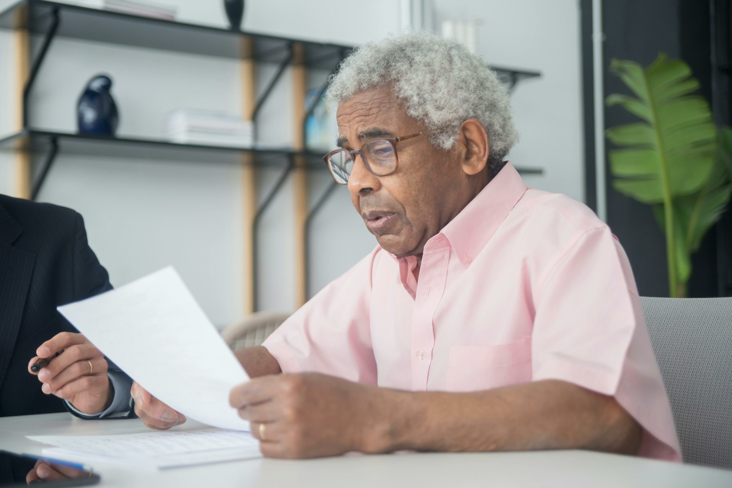 Elderly man with glasses reading a document in a home office setting, reflecting focus and concentration.